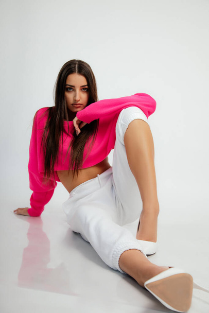 Gorgeous woman in pink crop pullover, pants and high heel shoes sitting on floor. Full length studio photo, white background. Young woman posing in sport casual wear. Fashion, stylish basic clothes. - 写真・画像