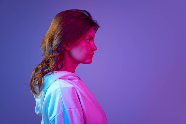 Side view portrait of young pretty girl looking straight with serious facial expression against gradient blue purple studio background in neon light. Concept of emotions, youth, lifestyle - 写真・画像