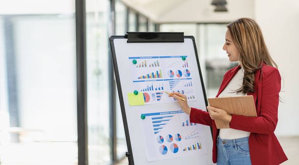 Asiatische Geschäftsfrau im Anzug steht lächelnd mit Stift und zeigt auf Dokumentdiagramm auf Whiteboard, das Daten im Konferenzraum präsentiert - Foto, Bild