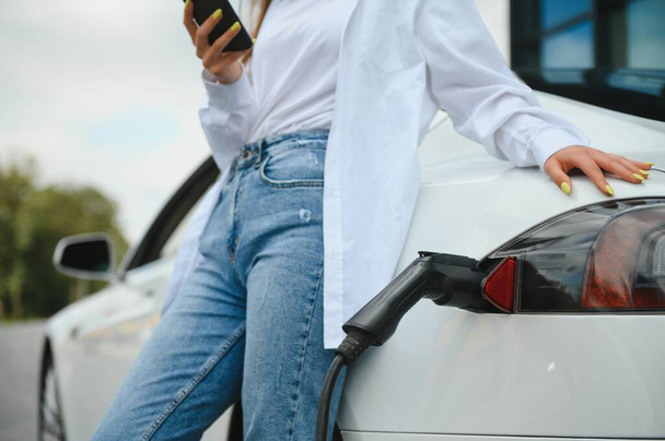 Phone in hands. Woman on the electric cars charge station at daytime. Brand new vehicle - Фото, зображення