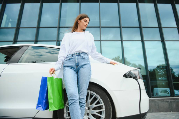 Happy young adult woman smiling wide, looking away, charging automobile battery from small public station, standing near electric car - Фото, изображение