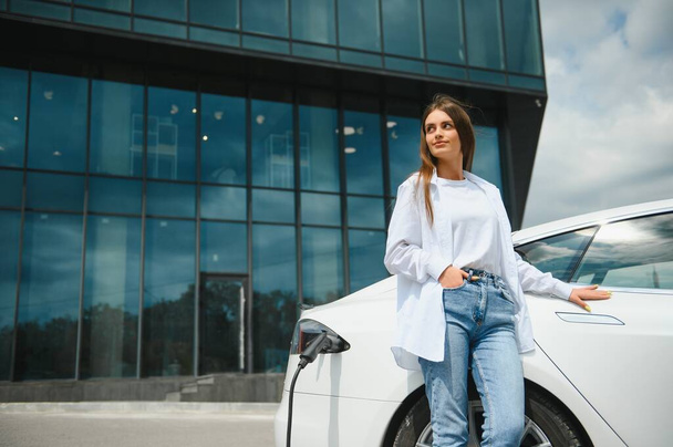 Happy young adult woman smiling wide, looking away, charging automobile battery from small public station, standing near electric car - Foto, imagen