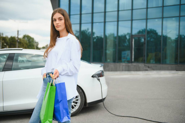 Happy young woman standing on city parking near electric car, charging automobile battery from small city station, holding shopping bags - 写真・画像