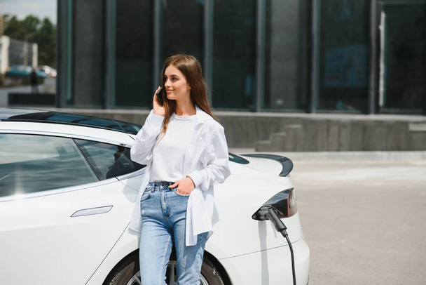 Happy young adult woman smiling wide, looking away, charging automobile battery from small public station, standing near electric car - 写真・画像