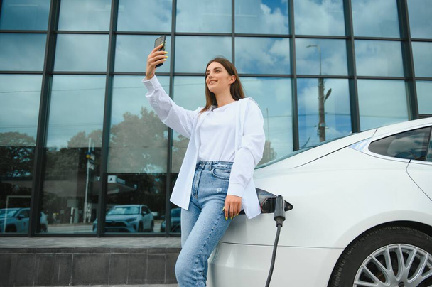 Woman with phone near an rental electric car. Vehicle charged at the charging station. - Foto, Imagen