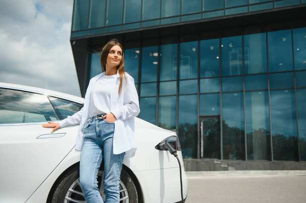 Woman near electric car. Vehicle charged at the charging station - Fotografie, Obrázek