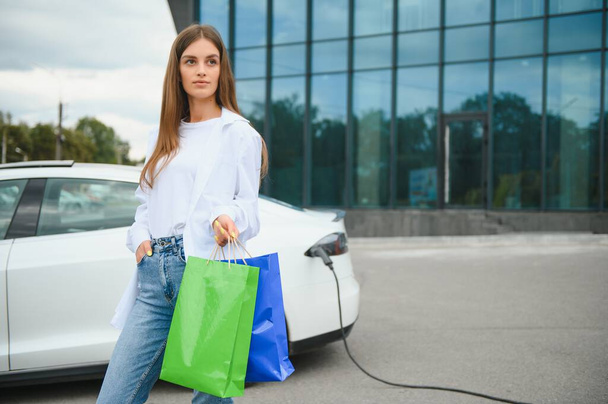 Happy young woman standing on city parking near electric car, charging automobile battery from small city station, holding shopping bags - Фото, изображение