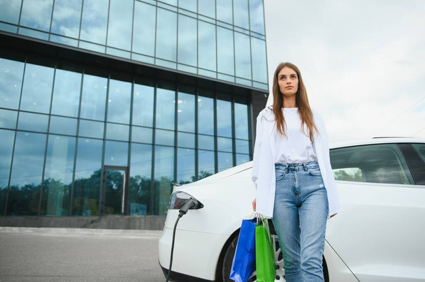 Happy young woman standing on city parking near electric car, charging automobile battery from small city station, holding shopping bags - Foto, Imagem