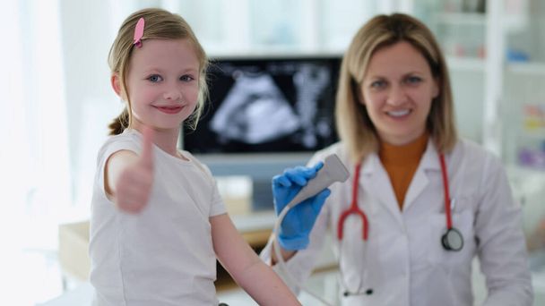 Маленькая девочка показывает большой палец на приеме у врача с ультразвуковой диагностикой в клинике. Высококачественное профессиональное инструментальное медицинское обследование в детской концепции - Фото, изображение