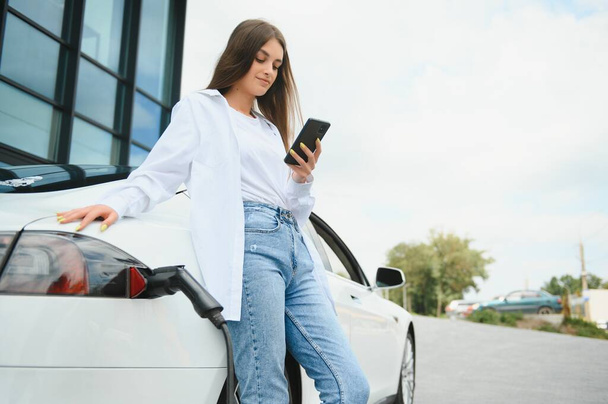 Phone in hands. Woman on the electric cars charge station at daytime. Brand new vehicle - Фото, изображение