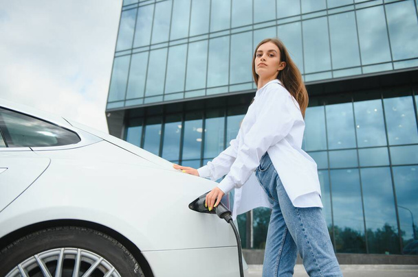 Smiling young caucasian woman plugging electricity cable in electric vehicle for charging on sunny mall parking, cropped. Lifestyle and ecology concept - 写真・画像