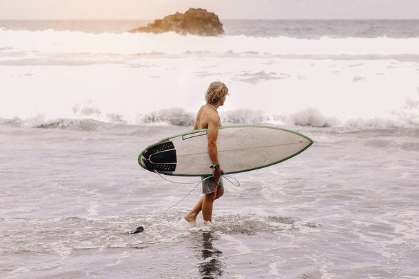 Back view of young fit surfer man with surfboard runs into ocean or sea with big waves for surfing. Concept of extreme sport, fitness, freedom, happiness, new modern life. Sports travel destination - Fotó, kép