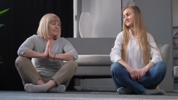 Nonna e figlia fanno yoga seduti sul pavimento a casa nel mezzo del soggiorno. Stile di vita sano - Filmati, video