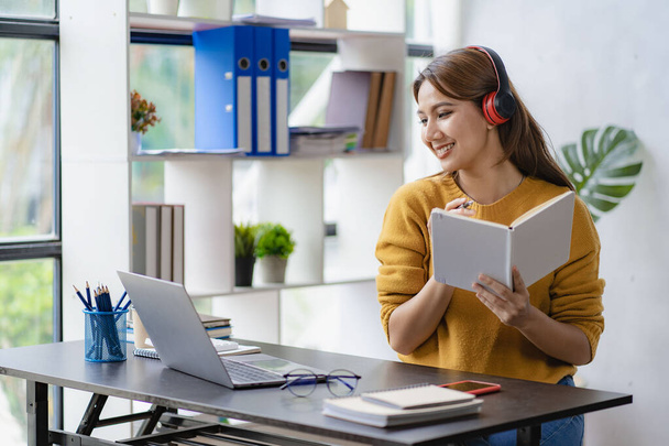Jovem estudante asiático em fones de ouvido e olhando para a câmera no futuro auto-educação ou conceito de aprendizagem, jovem estudante estudando on-line em casa com livro e laptop - Foto, Imagem