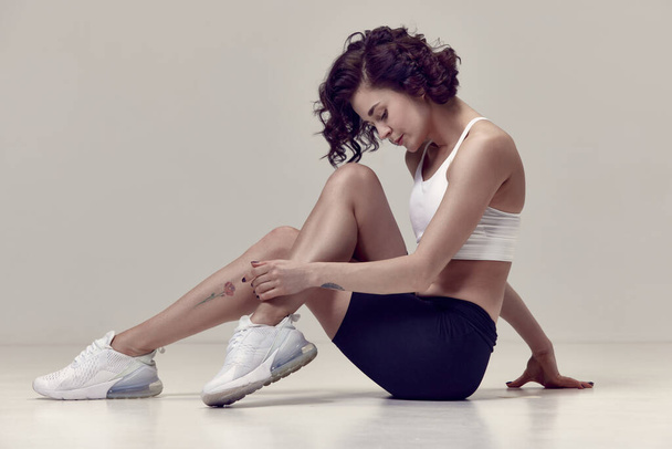 Young attractive woman with athletic, fit body, in sportswear sitting on floor, getting ready to train against grey studio background. Concept of sportive lifestyle, beauty, body care, fitness, health - 写真・画像