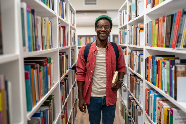 Lachend Afrikaans-Amerikaanse student die een boek vasthoudt in de universiteitsbibliotheek tussen de boekenplanken. Leven op de campus, onderwijs, zelfontwikkeling leesboeken studeren leren onderzoek concept. - Foto, afbeelding