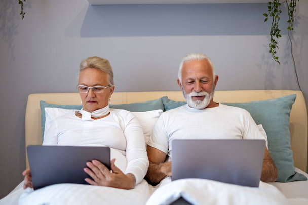 Happy senior couple using laptop while lying in bedroom.Technology and senior people concept. - Фото, изображение
