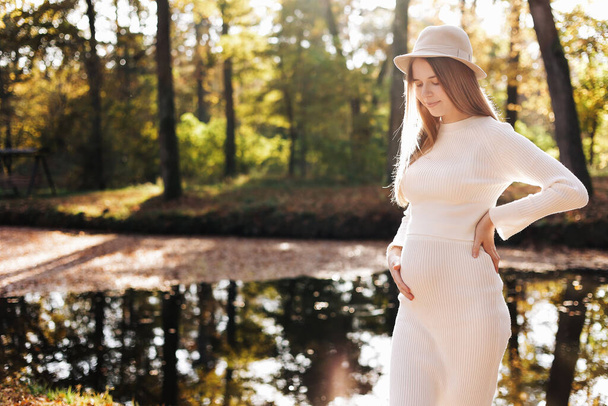 Ημέρα μητέρων. Όμορφη νεαρή έγκυος γυναίκα σε λευκό στενό πλεκτό φόρεμα με μικρή κοιλιά χαλαρώνοντας σε εξωτερικούς χώρους δίπλα στη λίμνη με αντανάκλαση της φύσης και του ήλιου στο πάρκο και κοιτάζοντας την κάμερα. Ημέρα γυναικών. - Φωτογραφία, εικόνα