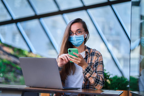 Portrait of woman freelancer in protective mask, wireless headphones and round eyeglasses using phone and computer for remote working online in cafe. Health protection in public places - Fotó, kép