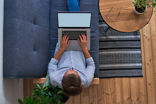 Freelancer mulher sentado no sofá e trabalhando remoto em um laptop em um confortável espaço de trabalho caseiro com plantas em ambientes fechados. Vista superior         - Foto, Imagem