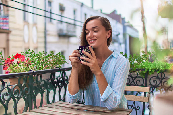 Beautiful happy smiling romantic woman with closed eyes enjoying of aromatic coffee on the balcony in the early morning in city - Zdjęcie, obraz