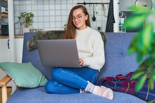 Усміхнена щаслива жінка в круглих окулярах і теплий білий светр сидить на дивані з ноутбуком вдома
        - Фото, зображення