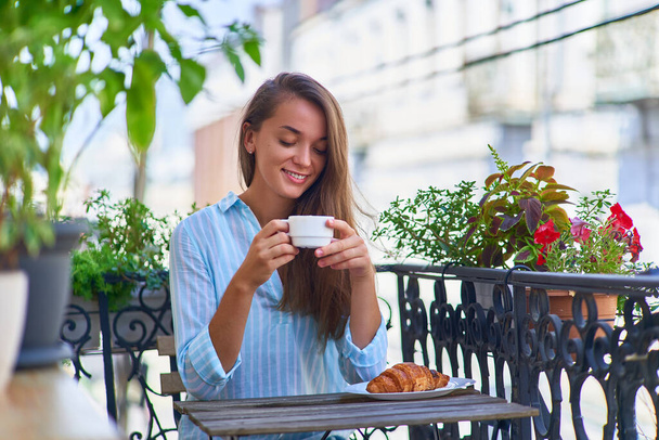 Portrait of a beautiful happy cute joyful smiling romantic woman with a aromatic coffee cup in hands and a plate of fresh baked croissant on the table in the morning on a balcony                      - Φωτογραφία, εικόνα