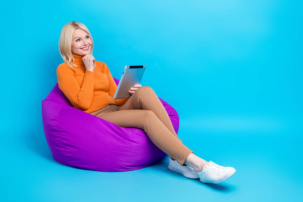 Full size photo of intelligent woman orange turtleneck sitting on pouf hold tablet look empty space isolated on blue color background. - Foto, Imagen