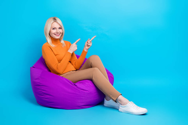 Full size photo of funny woman orange turtleneck sitting on pouf indicating at offer empty space isolated on blue color background. - Φωτογραφία, εικόνα