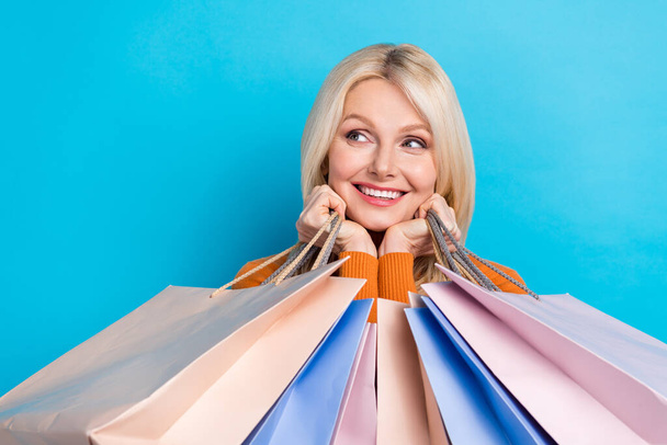 Portrait of lovely cheerful retired lady wear trendy clothes hold bags looking empty space special deal isolated on blue color background. - 写真・画像
