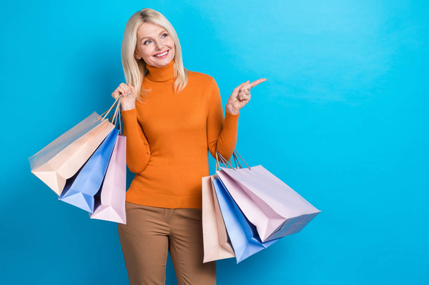 Photo of pensioner woman wear orange sweater bags shopaholic direct finger empty space new clothes brand isolated on cyan color background. - Φωτογραφία, εικόνα