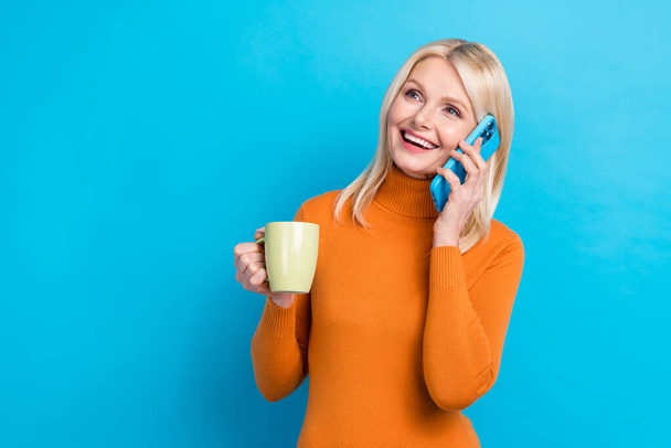 Portrait of cheerful retired lady wear orange trendy clothes speak telephone look empty space hold mug isolated on blue color background. - Fotografie, Obrázek