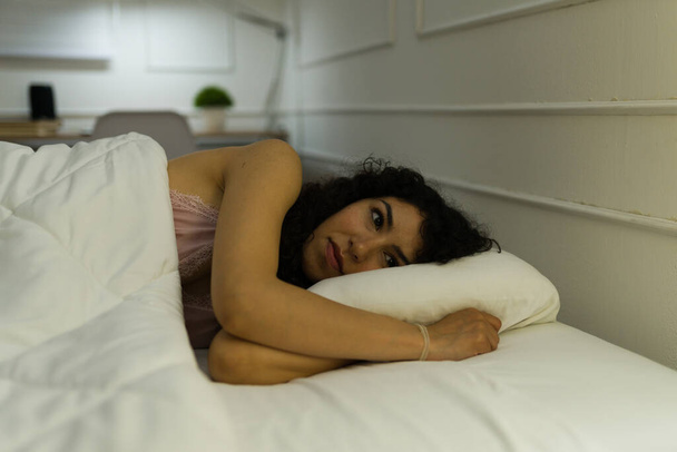 Krásná hispánská žena vypadá rozrušeně, zatímco trpí problémy se spaním a nespavostí pozdě v noci ve své ložnici - Fotografie, Obrázek