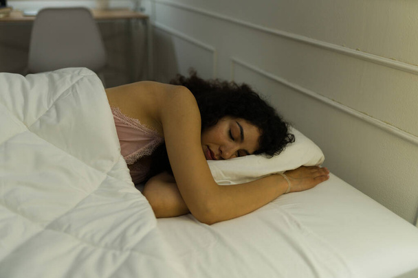 Красивая латинская женщина с вьющимися волосами выглядит расслабленной под одеялом в своей удобной кровати, спящей и отдыхающей  - Фото, изображение