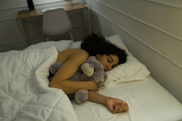 Szczęśliwa zrelaksowana kobieta przytula swojego misia śpiąc pod kołdrą w łóżku w nocy - Zdjęcie, obraz