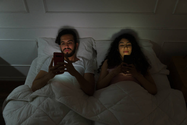 Pareja latina joven acostada en la cama mientras envían mensajes de texto en el teléfono inteligente o usan las redes sociales antes de dormir - Foto, Imagen