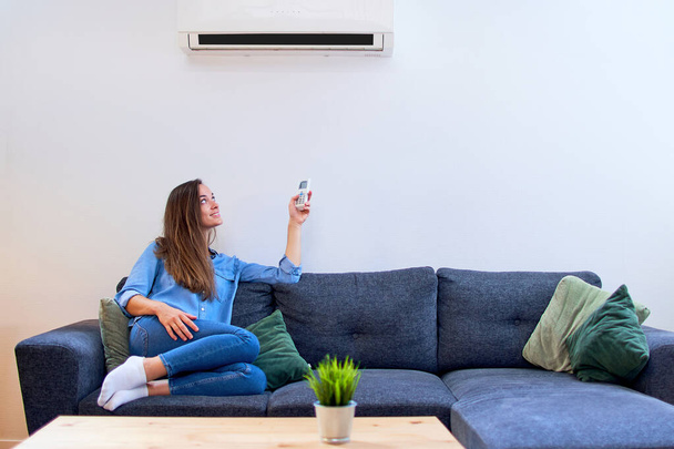 Young happy woman sitting on couch under air conditioner and adjusting comfort temperature with remote control at modern home - Foto, Bild