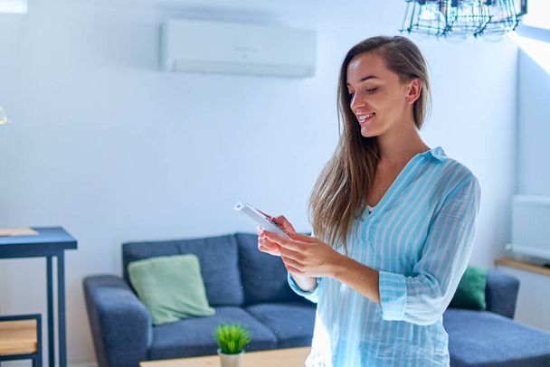 Young cute casual woman using air conditioner and adjusting comfortable temperature with remote control at apartment - Photo, image