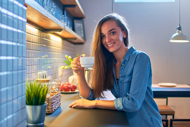 Portrait of beautiful smiling cute drinking woman holding a white cup and sitting at modern loft kitchen at home - Foto, imagen