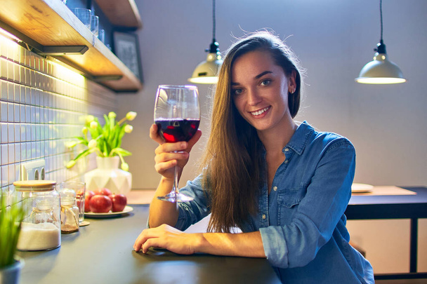Portrait of beautiful cute young casual smiling happy drinking woman holding a glass of red wine and sitting at the table in the loft style kitchen at home - Foto, Bild