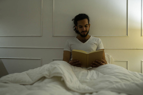 Chytrý mladý muž v pyžamu čtení knihy v posteli a vypadá uvolněně při odpočinku v ložnici před spaním - Fotografie, Obrázek