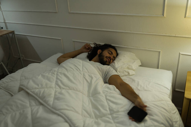 彼のスマートフォンを使用している間、ベッドの中で夜に眠りに落ちるカバーの下に疲れた若い男  - 写真・画像