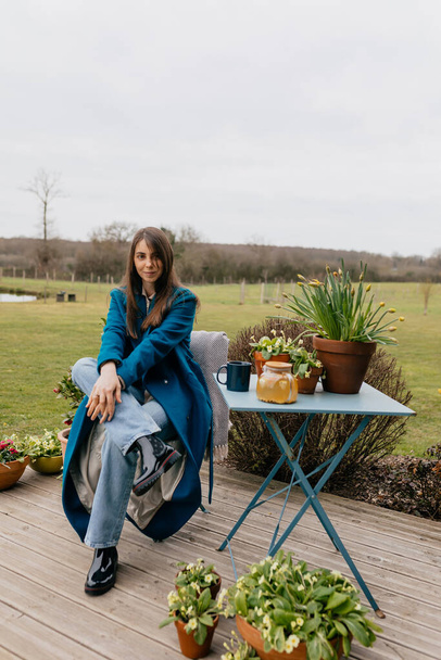 Plná délka venkovní fotografie stylové evropské ženy s dlouhými volnými vlasy na sobě dlouhý modrý kabát a džíny sedí na terase pod širým nebem s květinami proti zelené složené  - Fotografie, Obrázek