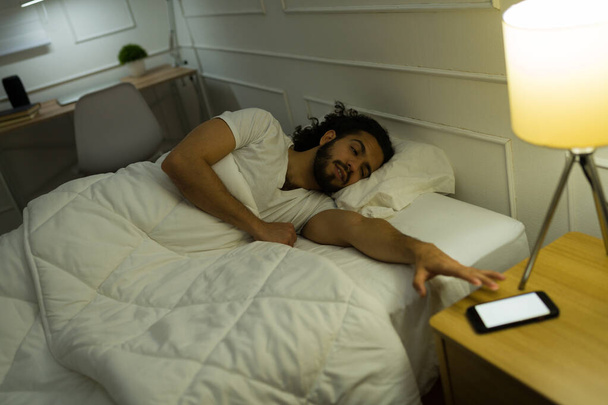 Fáradt fiatalember az ágyban felébred reggel, és kikapcsolja az ébresztőórát az okostelefonon  - Fotó, kép