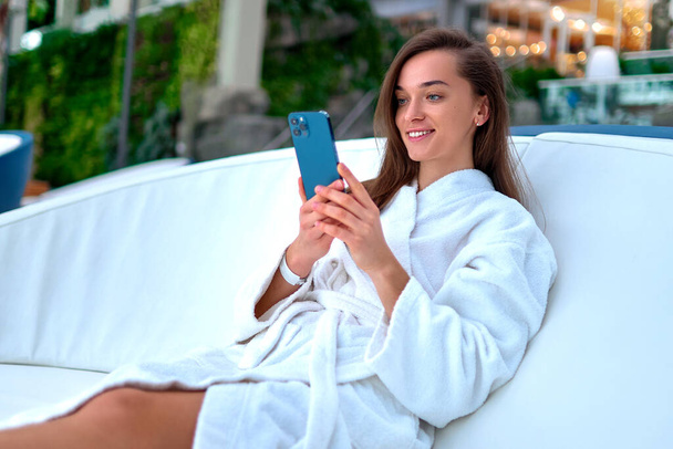 Young attractive smiling woman wearing white bathrobe using smartphone for watching video and browsing online while lying on a lounger during relaxing at spa resort               - Фото, изображение