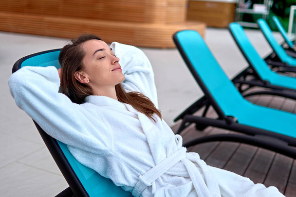 Calm serene woman wearing bathrobe with closed eyes and hands behind head relaxing and lying on a lounger at wellness spa resort. Satisfaction, feeling good and recreation time. Easy lifestyle - Fotó, kép