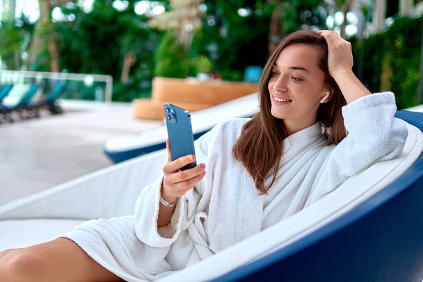 Mujer sonriente atractiva joven que usa albornoz blanco usando un teléfono inteligente para ver video y navegar en línea mientras está acostada en una tumbona durante la relajación en el balneario               - Foto, Imagen