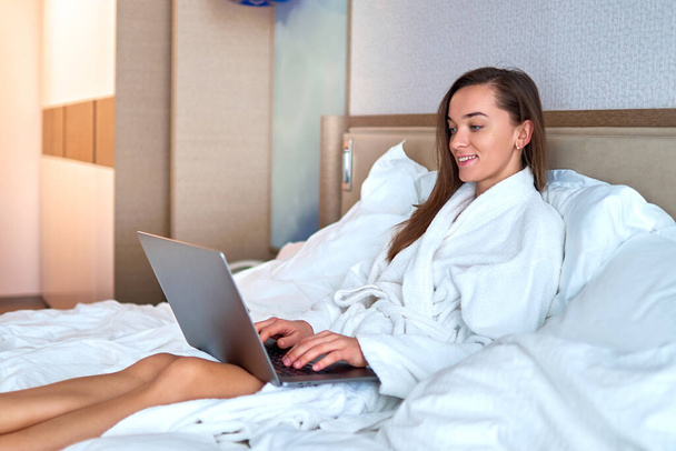 Młoda, urocza, uśmiechnięta kobieta freelancer w białym szlafroku zdalnie pracująca online przy komputerze na łóżku z pokoju hotelowego. Łatwy styl życia   - Zdjęcie, obraz
