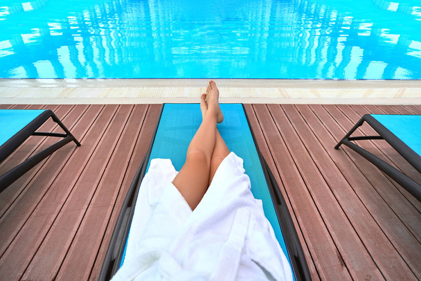 Жінка в халаті з красивими тонкими гладкими довгими ногами лежить на шезлонгу біля басейну, розслабляючись у оздоровчому курортному спа-центрі. Легкий спосіб життя і задоволення
 - Фото, зображення