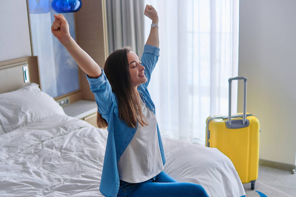 Satisfied joyful traveler woman with raised hands sitting on a bed in hotel bedroom after check-in and rejoicing travel start. Enjoy holiday and vacation. Happy moment - Photo, Image